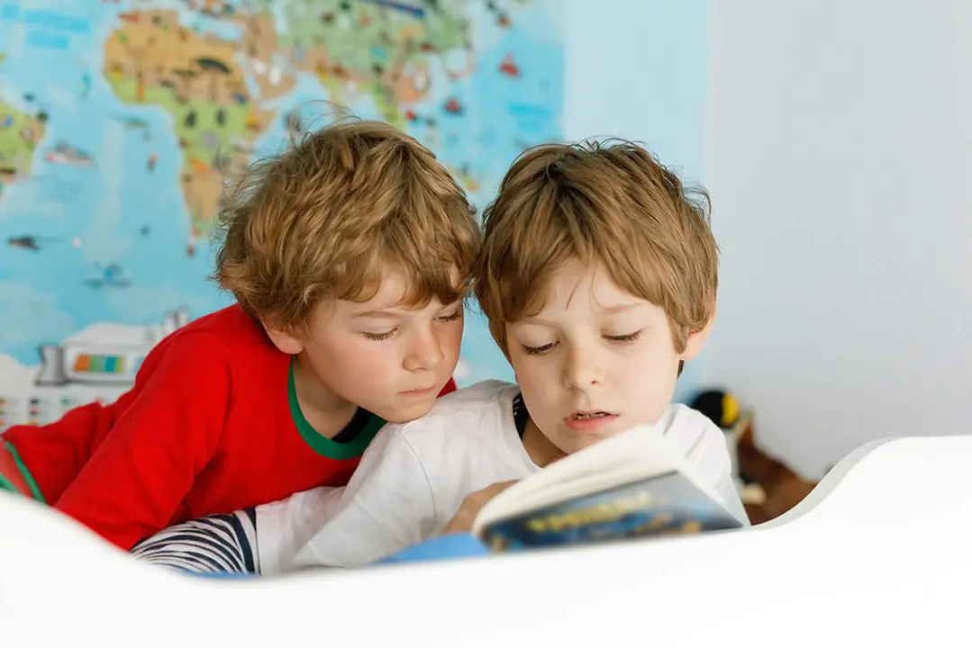  Two little kids boys in pajamas reading a book in bed. Excited siblings in nightwear. One brother boy reading loud and another child hearing. Family. 