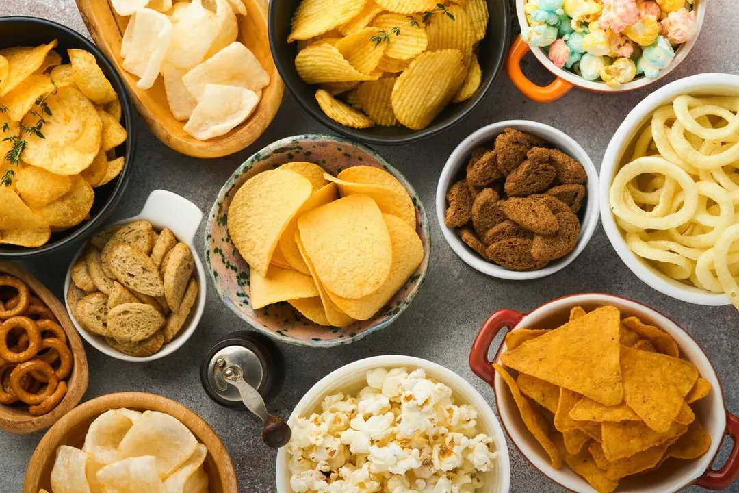 Snacks. Unhealthy food. All classic potato snacks with peanuts, popcorn and onion rings and salted pretzels in bowl plates on gray concrete background. Unhealthy food for figure, heart, skin, teeth