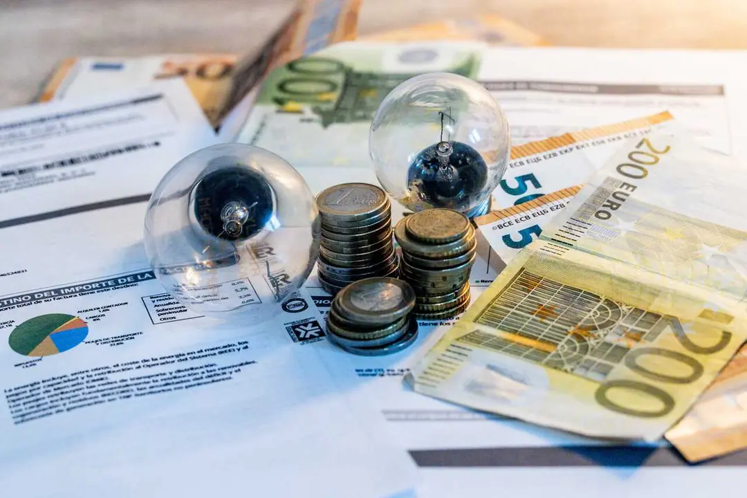 Archivo - Dos bombillas sobre billetes, monedas y facturas en imagen de archivo.