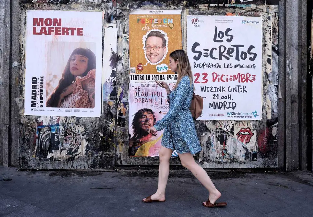 Archivo - Una mujer pasa con su tel&eacute;fono m&oacute;vil por varios carteles de espect&aacute;culos culturales y conciertos