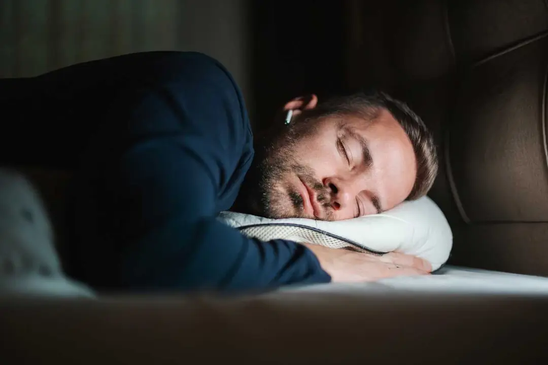 Young adult man sleeping soundly on a comfortable pillow in a dark bedroom, relaxing and recovering during the night while listening to music or podcasts through wireless earbuds
