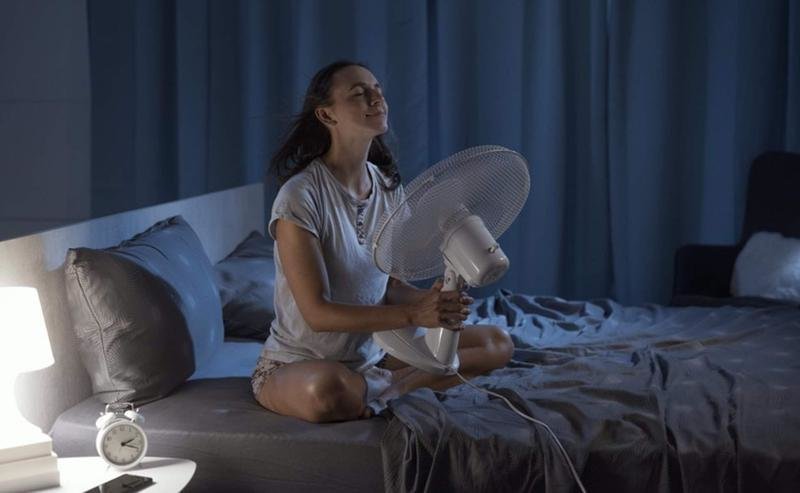 <p> Woman in her bedroom on a hot summer night, she is enjoying fresh air in front of a fan </p>