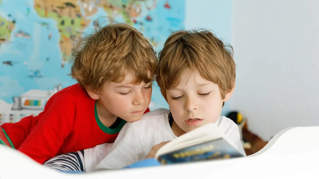  Two little kids boys in pajamas reading a book in bed. Excited siblings in nightwear. One brother boy reading loud and another child hearing. Family. 