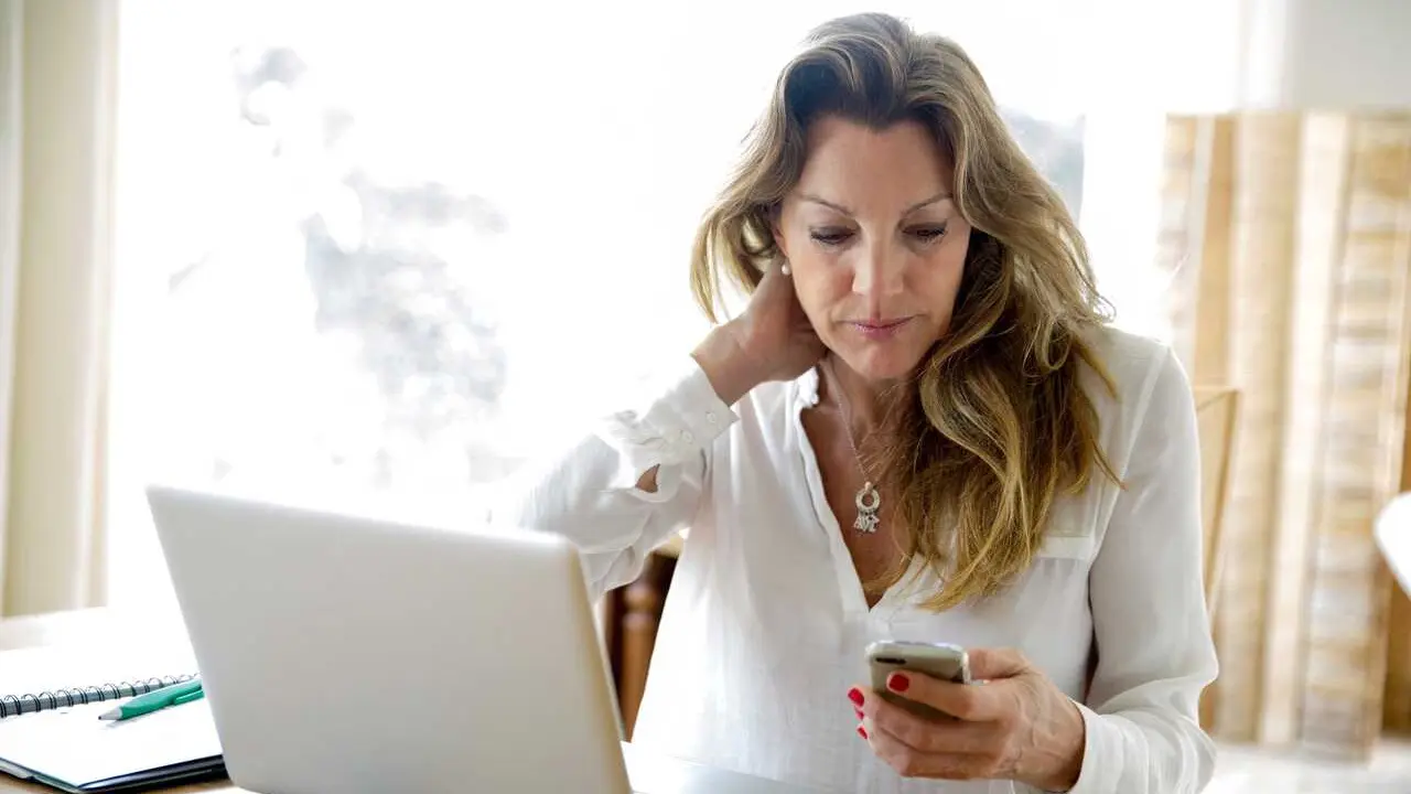 Archivo - Mujer frente al ordenador mientras consulta el m&oacute;vil