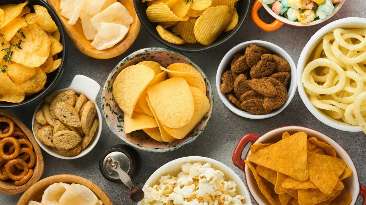 Snacks. Unhealthy food. All classic potato snacks with peanuts, popcorn and onion rings and salted pretzels in bowl plates on gray concrete background. Unhealthy food for figure, heart, skin, teeth