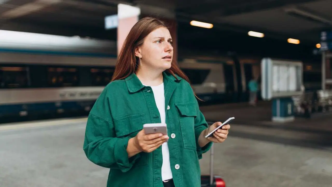 Young refugee redhhead woman waiting on station platform on background train, she lost and using smart phone. Railroad transport concept, Traveler.