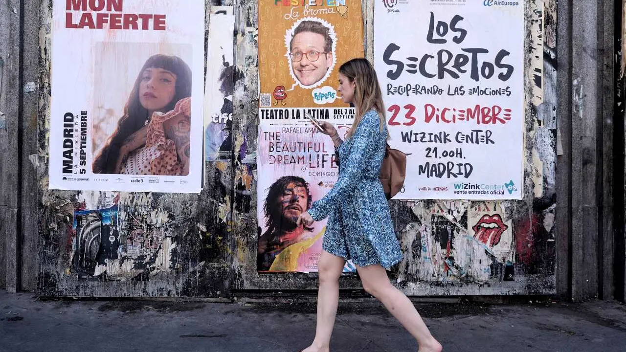 Archivo - Una mujer pasa con su tel&eacute;fono m&oacute;vil por varios carteles de espect&aacute;culos culturales y conciertos